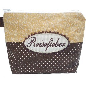 Puede incluir: Un neceser rectangular con cierre de cremallera. La mitad superior presenta un estampado floral en tonos beige y blanco, mientras que la mitad inferior es marrón con lunares blancos. Un parche ovalado bordado dice "Reiselieber".
