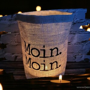 Puede incluir: Un portavelas de lino blanco con el texto "Moin. Moin." impreso en negro. El portavelas está encendido y rodeado de otras velas.