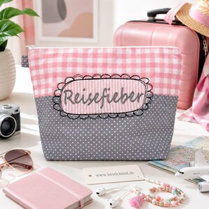 Könnte beinhalten: Eine Reißverschlusstasche mit einem oberen Bereich in rosa-weißem Vichy-Karo und einem unteren Bereich in Grau mit weißen Punkten. In der Mitte befindet sich ein bestickter, gewellter Aufnäher mit dem Wort Reisefieber in dunkelgrauem Garn.