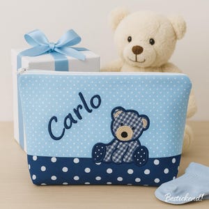 May include: A light blue cosmetic bag with white polka dots and the name "Carlo" embroidered in blue. A gingham teddy bear applique is on the front. A white gift box with a blue ribbon and a stuffed teddy bear are in the background.