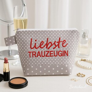 Könnte beinhalten: Eine graue Kosmetiktasche mit weißen Punkten und den rot gestickten Worten "liebste TRAUZEUGIN". Die Tasche hat einen Reißverschluss und einen kleinen Griff. Make-up und Schmuck sind um die Tasche herum angeordnet.