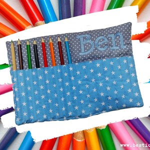 Op de afbeelding: Een blauwe etui met een wit sterrenpatroon en de naam "Ben" geborduurd in grijs. De etui is open en onthult verschillende kleurpotloden. De achtergrond toont een kleurrijke reeks potloden.