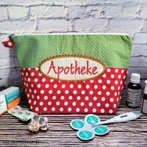 Puede incluir: Un neceser de lunares rojos y verdes con la palabra "Apotheke" bordada en la parte delantera. La bolsa tiene una cremallera blanca y un pequeño lazo rojo en el lateral. Varios suministros médicos están al lado de la bolsa.