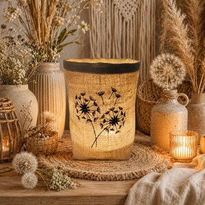 May include: A cylindrical burlap lantern featuring a dark brown rim and a black printed graphic of two dandelion seed heads. The textured fabric allows light to pass through, highlighting the intricate floral design on the front surface.