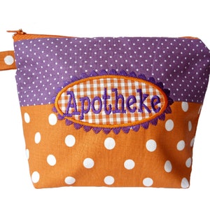 Puede incluir: Pequeño neceser con cremallera y diseño de bloques de color. La sección superior es morada con lunares blancos, y la inferior es naranja con lunares blancos. Un parche ovalado bordado en el centro dice "Apotheke".