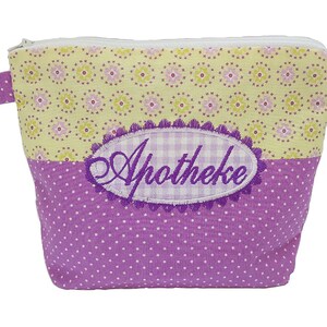 Puede incluir: Pequeño neceser rectangular con cierre de cremallera. La sección superior es de color amarillo claro con un estampado floral, la inferior es morada con lunares blancos, y el centro tiene un óvalo bordado con la palabra "Apotheke".
