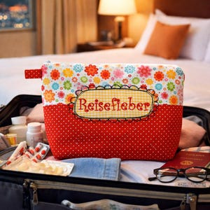 Puede incluir: Un estuche rectangular de tela con una sección superior de estampado floral colorido y una sección inferior roja con lunares blancos. Una etiqueta central de cuadros vichy amarillos muestra el texto rojo bordado "Reisefieber".