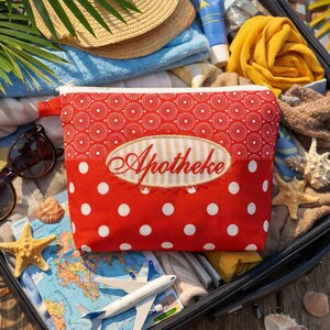 Puede incluir: Bolsa de cosméticos roja con lunares blancos con la palabra "Apotheke" bordada en la parte delantera. La bolsa está abierta, revelando artículos de viaje como un mapa, gafas de sol y un avión en miniatura. La bolsa está dentro de una maleta.