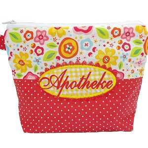 Puede incluir: Un estuche de tela con cremallera que presenta una sección superior con un colorido estampado floral y una sección inferior roja con lunares blancos. Un parche ovalado amarillo a cuadros está centrado con la palabra Apotheke.