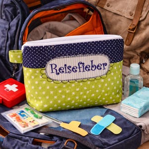 Puede incluir: Un neceser de viaje con cremallera blanca, con base verde con estrellas blancas y parte superior azul marino con lunares. La bolsa tiene la palabra "Reisefieber" bordada en la parte delantera. Otros accesorios de viaje son visibles.