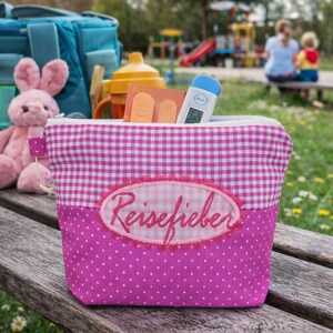 Può includere: Un astuccio da viaggio rosa e bianco a quadretti con la scritta "Reisefieber" ricamata sul davanti. L'astuccio contiene un termometro e altri oggetti. Un coniglietto di peluche rosa e uno zaino blu sono sullo sfondo.