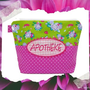 Puede incluir: Una bolsa de cremallera rosa y verde con lunares blancos y un parche bordado blanco que dice "Apotheke".