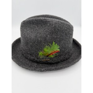 Pode incluir: Um chapéu fedora de feltro cinza-carvão com uma faixa trançada ao redor da base e um detalhe decorativo composto por penas verde-brilhante e laranja inseridas na lateral. O chapéu possui uma copa estruturada e aba larga e curvada.
