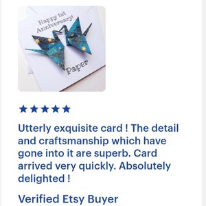 K&ouml;nnte beinhalten: Eine wei&szlig;e Gru&szlig;karte mit zwei Origami-Kranichen in Blau- und Goldt&ouml;nen. Die Karte tr&auml;gt die Aufschrift "Happy 1st Anniversary!" und "Paper". Die Kraniche sind aus gemustertem Papier gefaltet.