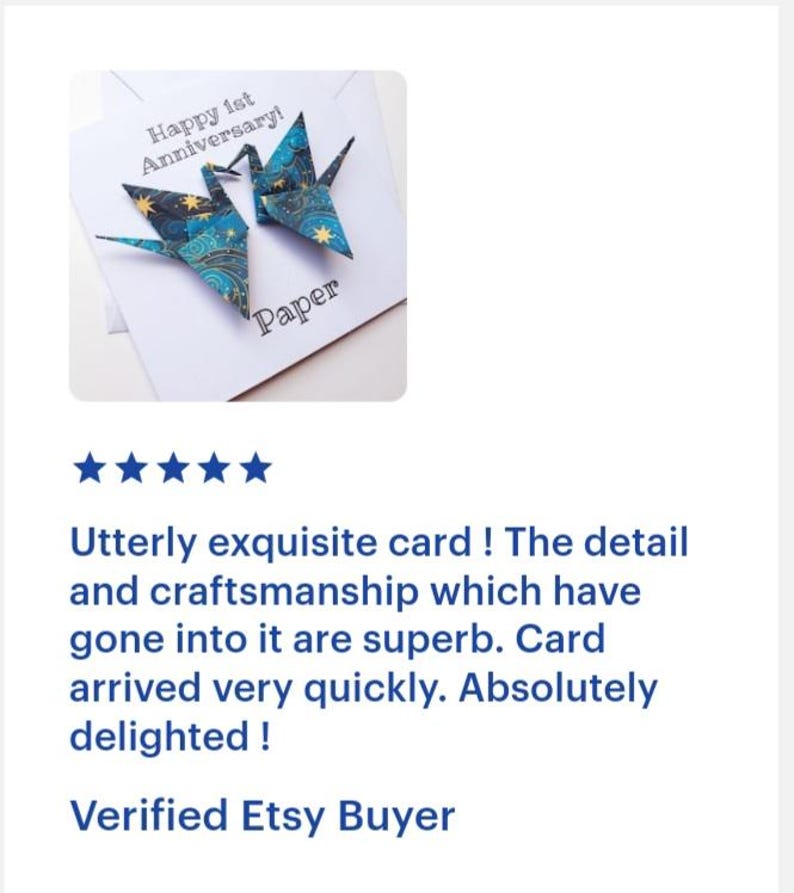 K&ouml;nnte beinhalten: Eine wei&szlig;e Gru&szlig;karte mit zwei Origami-Kranichen in Blau- und Goldt&ouml;nen. Die Karte tr&auml;gt die Aufschrift "Happy 1st Anniversary!" und "Paper". Die Kraniche sind aus gemustertem Papier gefaltet.