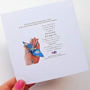 K&ouml;nnte beinhalten: Eine wei&szlig;e Karte mit Text &uuml;ber Origami-Papierkraniche und ihre Bedeutung in der japanischen Kultur. Ein blauer Origami-Kranich mit floralem Muster wird in einer Hand gehalten. Der Text erw&auml;hnt Gl&uuml;ck und langes Leben.