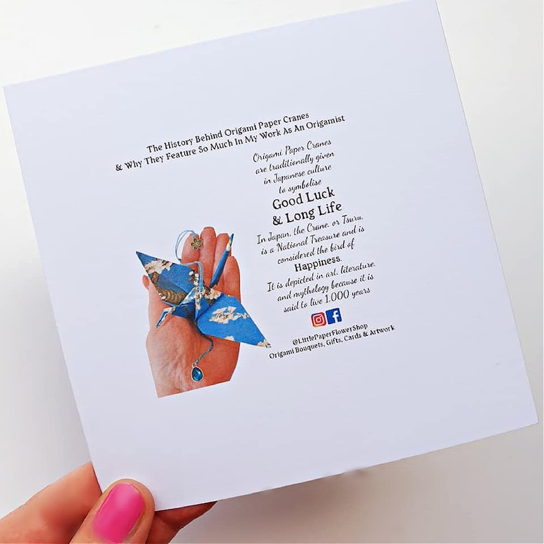 K&ouml;nnte beinhalten: Eine wei&szlig;e Karte mit Text &uuml;ber Origami-Papierkraniche und ihre Bedeutung in der japanischen Kultur. Ein blauer Origami-Kranich mit floralem Muster wird in einer Hand gehalten. Der Text erw&auml;hnt Gl&uuml;ck und langes Leben.