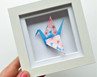 Könnte beinhalten: Ein quadratischer weißer Rahmen mit einem gefalteten Origami-Kranich aus Papier. Der Kranich besteht aus blauem Papier mit einem wiederkehrenden Muster aus hellrosa Kirschblüten mit dunkelrosa Zentren. Der Rahmen hat einen weißen Rand.