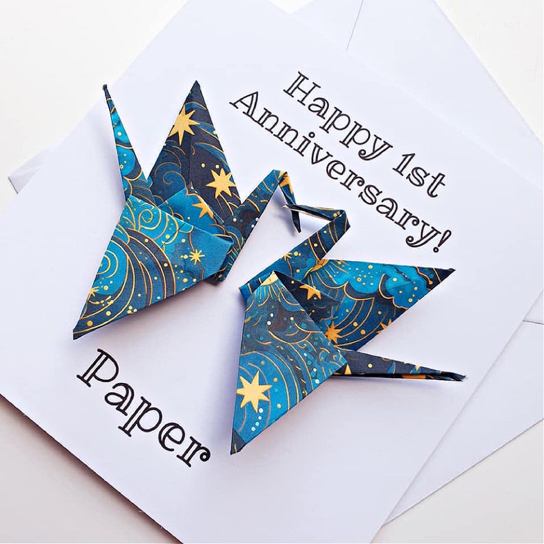 K&ouml;nnte beinhalten: Eine wei&szlig;e Karte mit dem Text "Happy 1st Anniversary!" und "Paper". Zwei Origami-Kraniche mit einem blauen, goldenen und wei&szlig;en Himmelsmuster befinden sich auf der Karte.