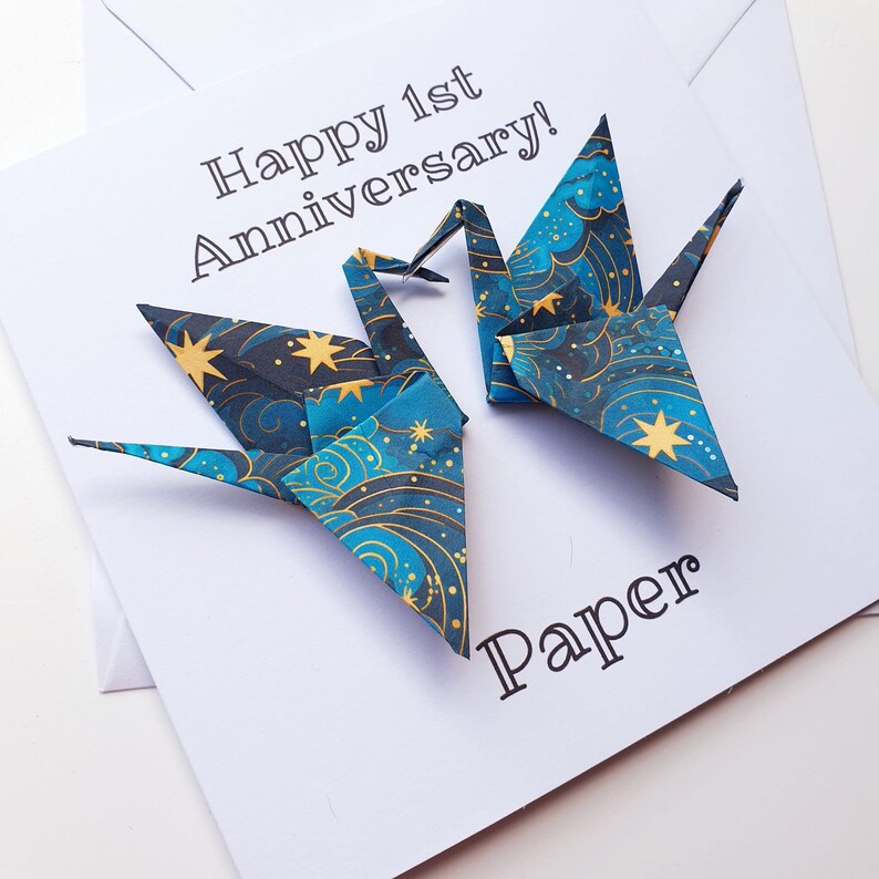 K&ouml;nnte beinhalten: Eine wei&szlig;e Karte mit dem Text "Happy 1st Anniversary!" und "Paper". Zwei Origami-Kraniche mit einem blauen, goldenen und wei&szlig;en Himmelsmuster befinden sich auf der Karte. Die Karte liegt auf einem wei&szlig;en Umschlag.
