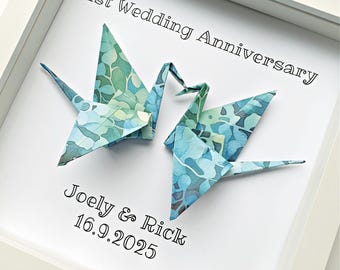 Könnte beinhalten: Ein weiß gerahmtes Wandbild mit zwei Origami-Kranichen aus blau-grün gemustertem Papier. Der Text lautet 1st Wedding Anniversary, Joely & Rick und 16.9.2025. Die Kraniche sind mittig vor einem schlichten weißen Hintergrund angeordnet.