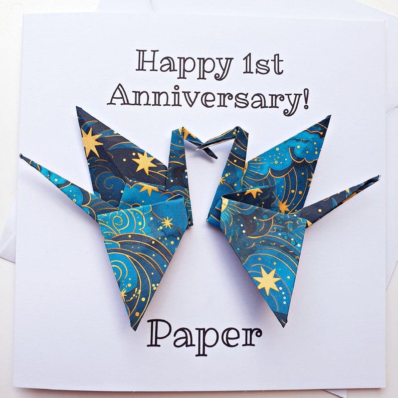 K&ouml;nnte beinhalten: Eine wei&szlig;e Karte mit dem Text "Happy 1st Anniversary!" und "Paper". Auf der Karte sind zwei Origami-Kraniche mit einem blauen und goldenen Himmelsmuster abgebildet. Die Kraniche sind herzf&ouml;rmig gefaltet.