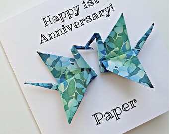 Könnte beinhalten: Eine weiße Karte mit dem Text "Happy 1st Anniversary!" und "Paper". Zwei Origami-Kraniche in Blau- und Grüntönen sind abgebildet. Die Kraniche haben ein Blumenmuster.