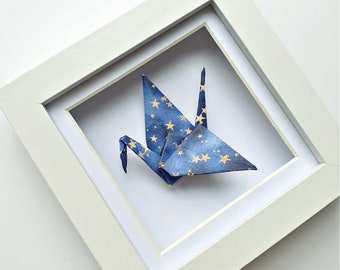 Könnte beinhalten: Ein gerahmter Origami-Kranich. Der Kranich ist aus blauem Papier mit einem goldenen Sternmuster gefaltet. Der Rahmen ist weiß und quadratisch, mit einem weißen Passepartout. Das Kunstwerk wird auf weißem Hintergrund präsentiert.
