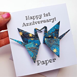 K&ouml;nnte beinhalten: Eine wei&szlig;e Karte mit dem Text "Happy 1st Anniversary!" und "Paper". Zwei Origami-Kraniche mit blauem und goldenem Himmelsmuster sind abgebildet. Die Karte wird von einer Hand mit rosa Nagellack gehalten.