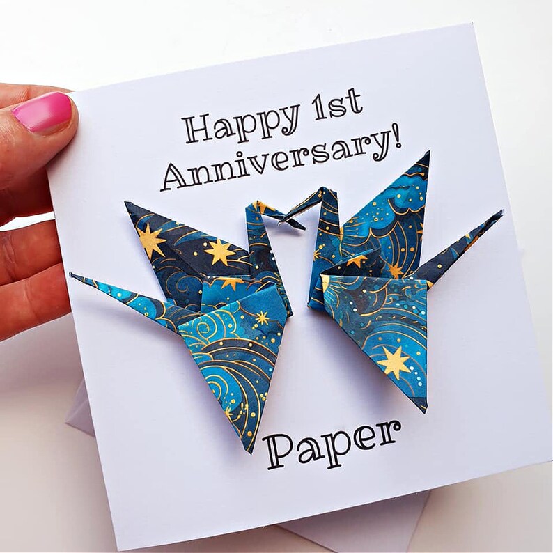 K&ouml;nnte beinhalten: Eine wei&szlig;e Karte mit dem Text "Happy 1st Anniversary!" und "Paper". Zwei Origami-Kraniche mit blauem und goldenem Himmelsmuster sind abgebildet. Die Karte wird von einer Hand mit rosa Nagellack gehalten.