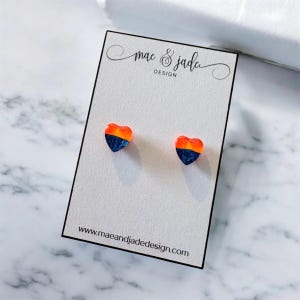 May include: A pair of heart-shaped stud earrings featuring a two-tone design with a vibrant orange top half and a deep navy blue bottom half. The earrings are displayed on a white card printed with the text mae & jade design and the website address.