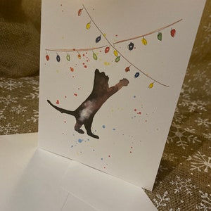 May include: A white greeting card with a watercolor illustration of a brown cat reaching for colorful Christmas lights. The card is accompanied by a white envelope. Splatters of paint in various colors add to the festive design.
