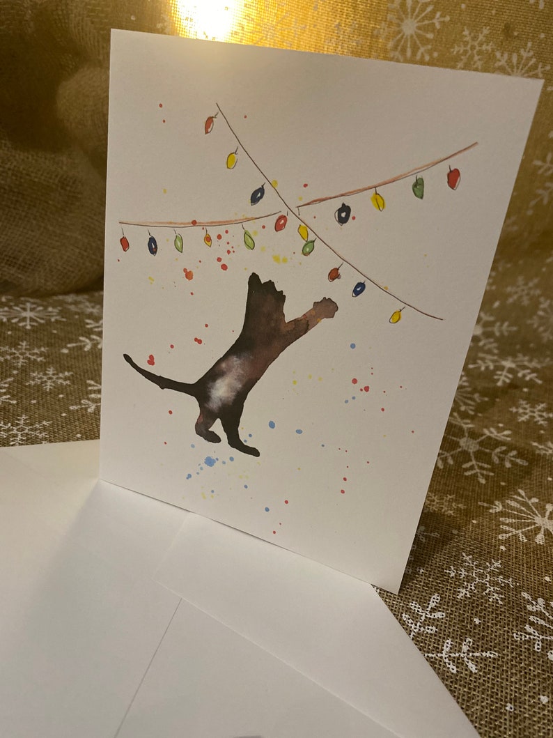 May include: A white greeting card with a watercolor illustration of a brown cat reaching for colorful Christmas lights. The card is accompanied by a white envelope. Splatters of paint in various colors add to the festive design.