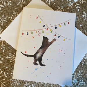 May include: Watercolor greeting card featuring a brown cat reaching for a string of colorful Christmas lights. The cat is depicted with watercolor paint splatters. Includes a white envelope. Holiday theme.
