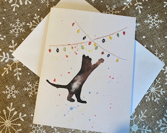 Op de afbeelding: Waterverf wenskaart met een bruine kat die naar een slinger van kleurrijke kerstverlichting reikt. De kat is afgebeeld met aquarelverf spetters. Inclusief een witte envelop. Feestdagen thema.