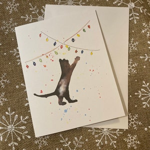 May include: A watercolor greeting card featuring a brown cat reaching for colorful Christmas lights. The card has a white background with colorful paint splatters. An envelope is included.