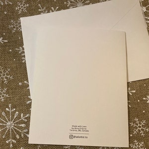 May include: A white greeting card with a matching envelope. The card has text that reads "Made with Love by Anna D Kuras Toronto, ON, Canada" and an Instagram handle. The card is on a textured surface.
