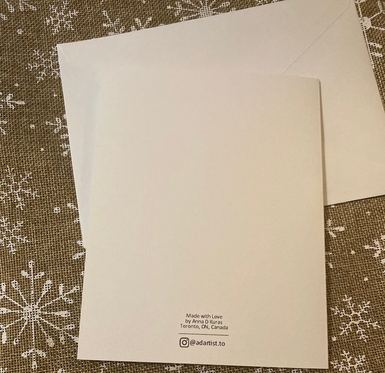 May include: A white greeting card with a matching envelope. The card has text that reads "Made with Love by Anna D Kuras Toronto, ON, Canada" and an Instagram handle. The card is on a textured surface.