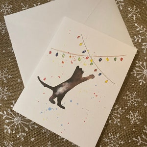 May include: A greeting card with a watercolor illustration of a dark cat reaching for colorful Christmas lights. The card is paired with a white envelope. Splatters of paint in red, blue, and yellow add to the festive design.
