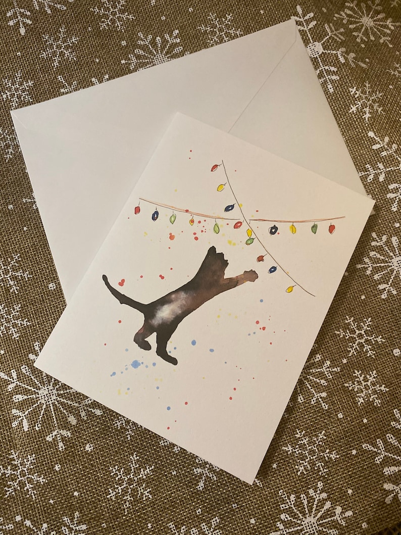 May include: A greeting card with a watercolor illustration of a dark cat reaching for colorful Christmas lights. The card is paired with a white envelope. Splatters of paint in red, blue, and yellow add to the festive design.