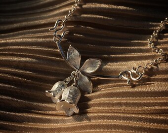 May include: A silver necklace with a delicate flower design and three bell-shaped charms hanging from the stem. The necklace is on a brown fabric background.