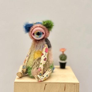 Pode incluir: Uma criatura de pelúcia caprichosa e colorida com um olho grande e pele multicolorida senta-se em uma caixa de madeira. A pelúcia tem braços e pernas estampados, e um pequeno cacto em um vaso preto está ao lado. O olho da criatura é azul e a pele é uma mistura de rosa, azul, verde e amarelo.
