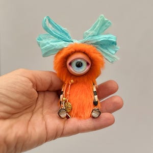 May include: A whimsical, handmade orange plush creature with a large, detailed eye. It has a blue bow, small feet, and striped arms. The creature has small gold charms hanging from its neck. A unique, artistic doll.