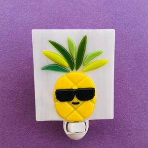 May include: A novelty night light featuring a yellow pineapple wearing black sunglasses on a white background. The pineapple has green and yellow leaves. The night light has a white on/off switch and is set against a purple background.