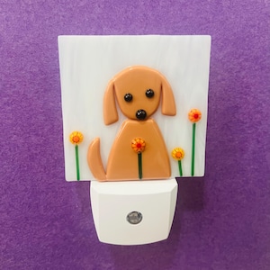 May include: A decorative night light featuring a brown dog with black eyes and a flower. The dog is set against a white background with orange and yellow flowers. The base of the night light is white.