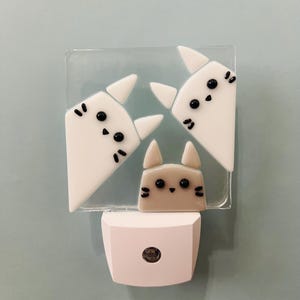 Puede incluir: Luz de noche decorativa con diseño de gato. La luz tiene una base blanca y una parte superior rectangular transparente con tres caras de gato en blanco y gris. Los gatos tienen ojos y bigotes negros.