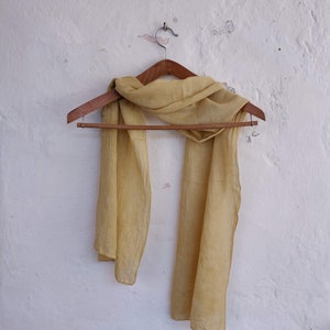 May include: A light yellow scarf hangs on a wooden hanger against a white wall. The scarf is made of a lightweight fabric and drapes loosely. The wall has a textured, distressed appearance.