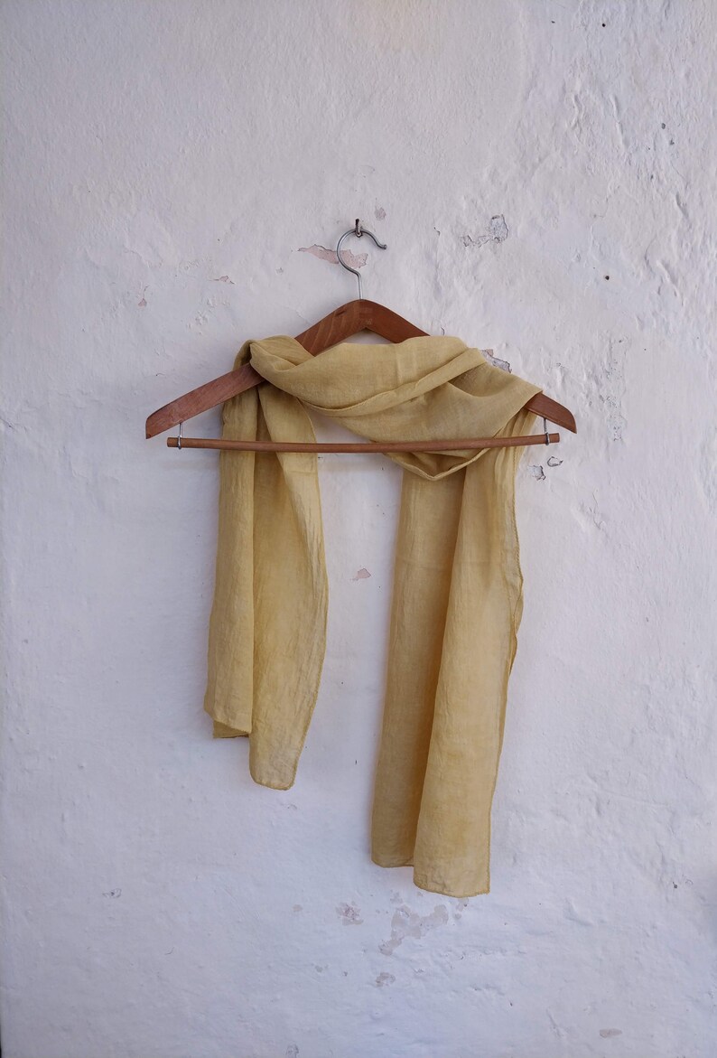 May include: A light yellow scarf hangs on a wooden hanger against a white wall. The scarf is made of a lightweight fabric and drapes loosely. The wall has a textured, distressed appearance.