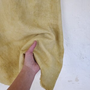 May include: A piece of yellow-green fabric with a textured appearance. The fabric is held up against a white wall, showing its natural, slightly uneven texture and a frayed edge. The fabric has a faded, aged look.