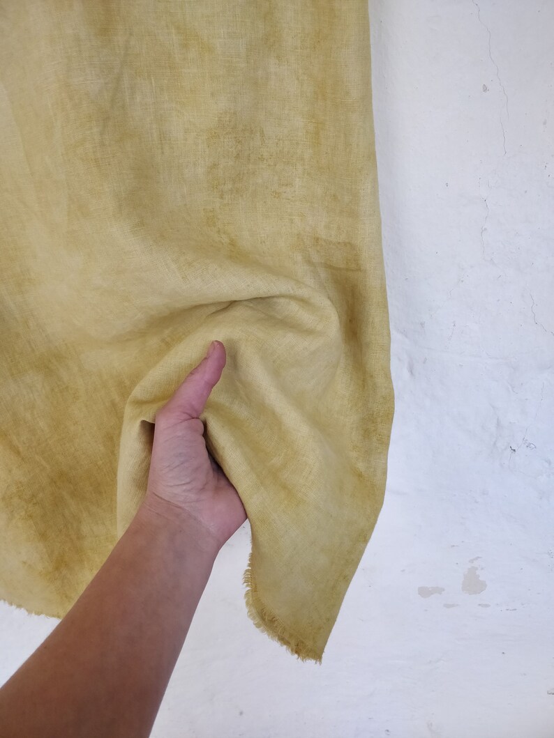 May include: A piece of yellow-green fabric with a textured appearance. The fabric is held up against a white wall, showing its natural, slightly uneven texture and a frayed edge. The fabric has a faded, aged look.
